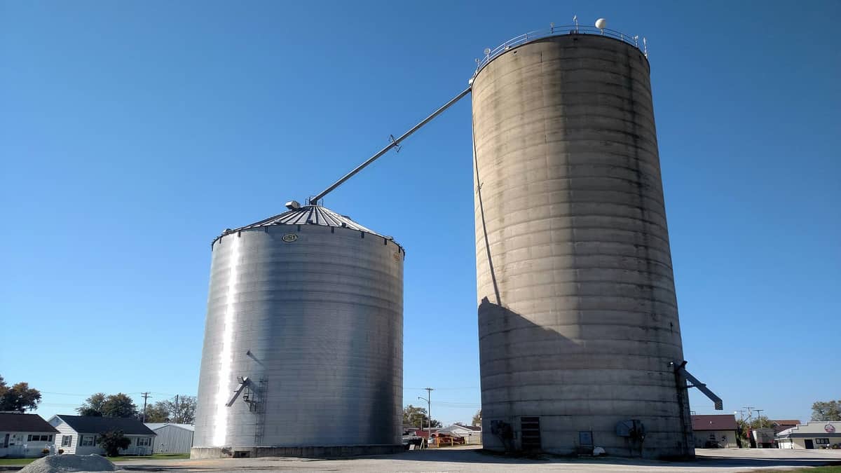 Dois silos de armazenagem de grãos, um metálico e outro de concreto, sob céu azul. Imagem ilustrativa